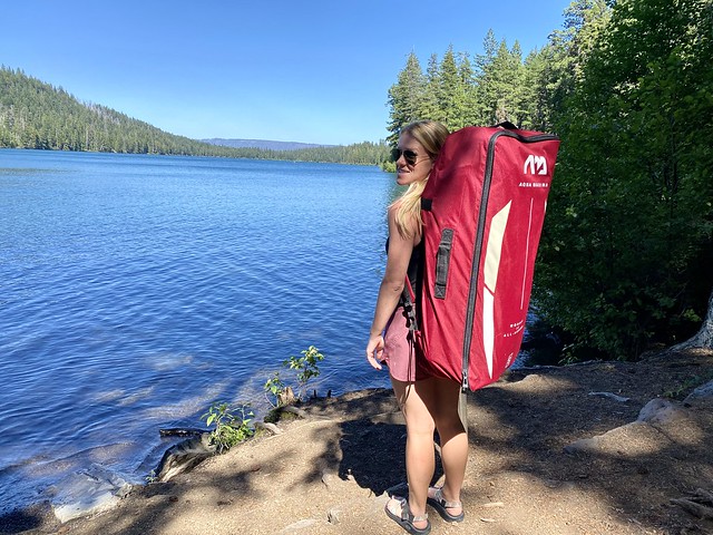 Kendall Brown is 5' 2" tall and carries her Aqua Marina SUP, with carrying case, from the car to the beach by herself. She is standing with her paddleboard, before assembled, at Suttle Lake in Central Oregon.