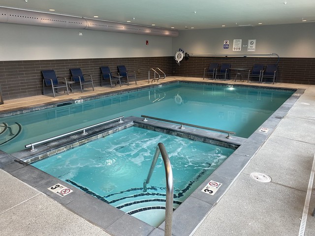 Best Western Premier Peppertree rectangular, indoor swimming pool. A whirlpool tub sits squarely within the large hotel swimming pool. A metal handrail and four steps lead into the whirlpool tub. Four metal chairs are to the left of the indoor swimming pool. 