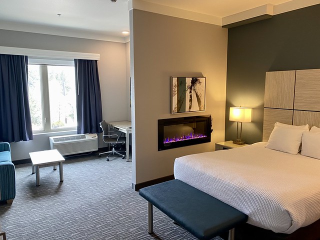 Best Western queen bed with gas fireplace mounted on the wall. A nature photo is above the rectangular gas fireplace. A blue, fabric-covered, bench is at the foot of the queen bed. A large window with blue drapes is beyond the queen bed. A small desk with chair and heating and air conditioning unit is mounted by the hotel window. 