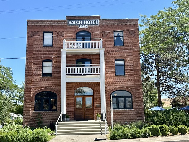 Three-story, red brick, historic Balch Hotel with white block letters on the top of the building, reading Balch Hotel 1907. Rectangular shaped glass windows are on either side of the top 2 stories of the hotel. Arcade-shaped windows are in the center of the brick building on top 2 stories. Ground floor glass door front entrance to the hotel is framed with 2 white banisters, serving as the base of the top two balconies over the 2nd and 3rd floor. Five cement steps lead to the front porch hotel entrance. 