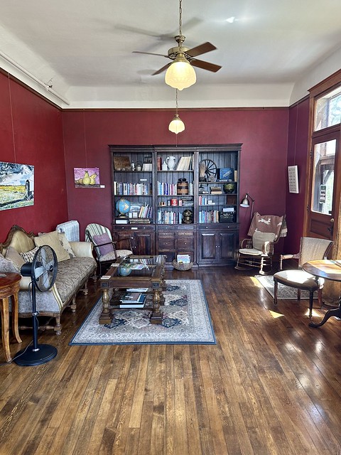Balch Hotel parlor with bookcase. Brick red colored walls feature local art. A wooden antique bookcase holds books, a globe and antique black manual typewriter. A wooden rocking chair is to the right of the bookcase. Antique sofa is to the left of the room with small, oriental rectangular rug over the dark, hardwood plank floors. A small, portable floor fan is to the right of the sofa. 
