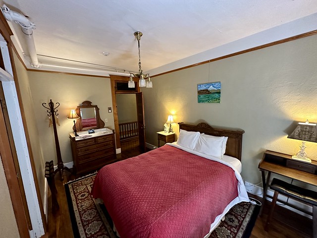 Queen bed with red brick colored bedspread. Three-pronged antique lamp suspended from ceiling. Square Oriental rug is under queen bed, over dark hardwood floors. A small Mt. Hood acrylic painting hangs over the bed and wooden headboard. 