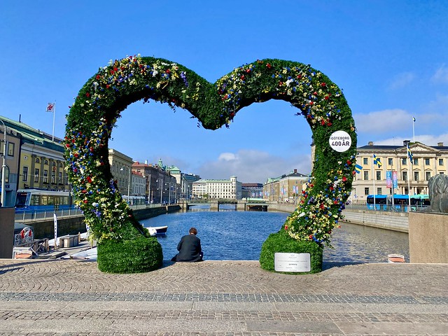 Heart-shaped floral greenery at Brunnsparkbron in Gothenburg waterway. A man is sitting with his back to the camera in the left bottom of the heart art structure. 