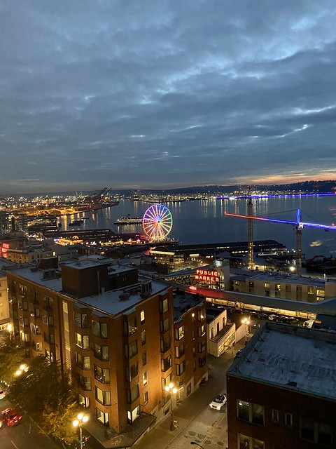 Ferris wheel at night with multi-colored lights. Puget sounds in the distance and Pike Place Market sign in the foreground. Stewart Street and single white car below. 