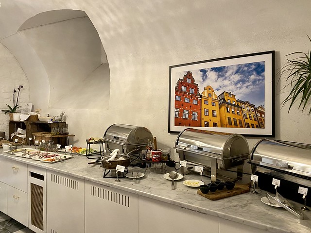 Breakfast buffet at Hotel Gamla Stan features hot and cold items, including scrambled eggs, bacon, swedish meatballs, quiche, plates of ham, turkey, brie and blue cheese, havarti cheese, hard boiled eggs and baskets of bread. A photo of Stockholm buildings hangs above the breakfast buffet silver warming trays. 