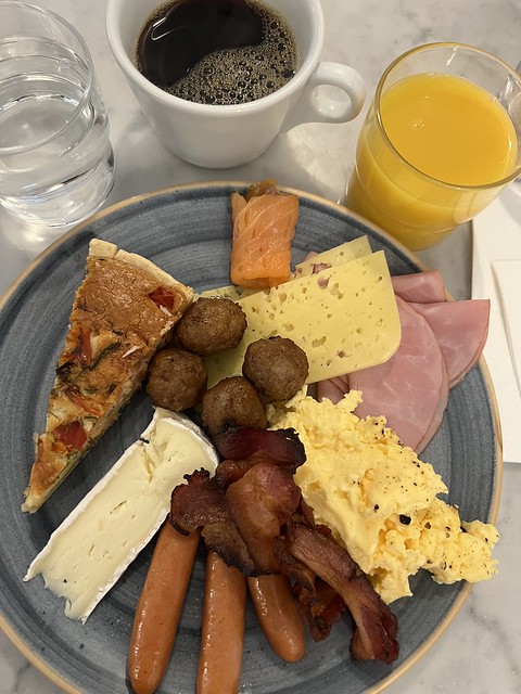 Breakfast plate at Hotel Gamla Stan includes sausage, bacon, Swedish meatballs, sliced ham, Havarti cheese, scrambled eggs, brie cheese wedge, small slice quiche and piece of smoked salmon. Glass of water, cup of coffee and glass of orange juice are top of blue plate. 