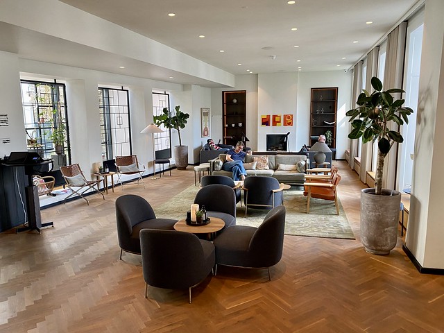 Hotel Kong Arthur lobby with parquet wood floor and white painted walls. Two large potted fig trees sit on either side of the hotel lobby. Four dark brown chairs of Danish design, surround a small, circular wood table. A white candle is on the table. A large beige, cloth sofa is behind the 4 chairs. Sofa and 2 additional chairs are on top of olive-colored, square area rug. Three small, square orange, yellow and red paintings are on the back, white wall. 