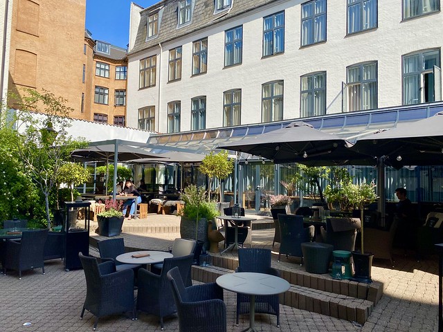 Hotel Kong Arthur patio sits in the courtyard of the Copenhagen hotel. Circular small bistro-style tables are situated in the courtyard with black wicker chairs for hotel guests to relax or eat breakfast outdoors. Potted trees are situated throughout the courtyard. 