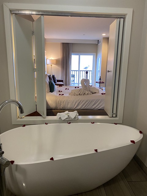 Large white, freestanding soaking bathtub with peek-a-boo shutter wooden windows to king bed and ocean view. 