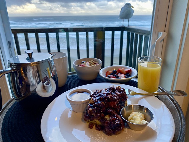 Complimentary breakfast included with hotel stay. Marrionberry French toast, fresh strawberries and blueberries, yogurt, granola and fruit, coffee and orange juice. Seagull on hotel balcony looks on with ocean in distance. 