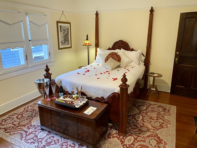 Lady Florence suite with rose petals scattered on queen bed. A romance package of sparkling wine, chocolate charcuterie board and 2 champagne flutes are on a wooden hope chest at the base of the queen bed. 