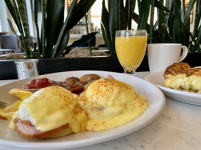 Eggs Benedict with roasted potatoes and a separate plate of quiche, are accompanied with a glass of orange juice and mug of coffee on a white marble table top at 5th Street Bistro restaurant inside Majestic Inn hotel. 