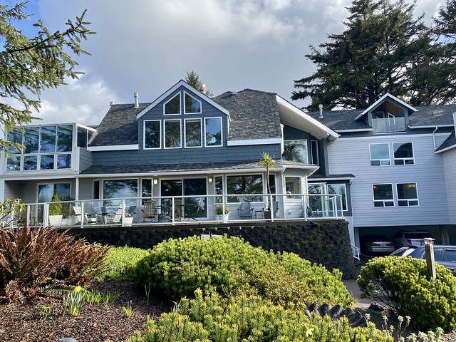 Three-story Ocean House in Newport, Oregon. Cap Cod style beach house with blue and white wood siding and glass windows facing Agate Beach. Large patio on rear of the oceanfront inn has Adirondack chairs on cement patio. Green shrubs surround the patio, from below. 