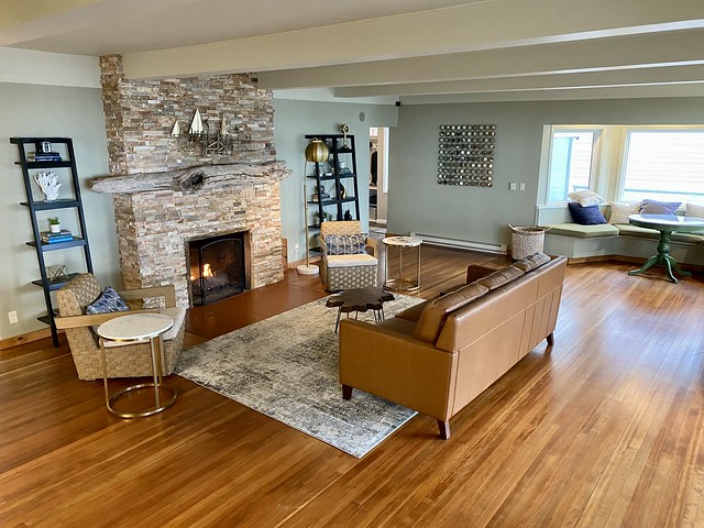 Ocean House living room with gas fireplace and stone, floor to ceiling, wall. Beige leather sofa, small, burl slab table and two beige, wicker chairs are in front of the fireplace, placed on a rectangular area rug. 