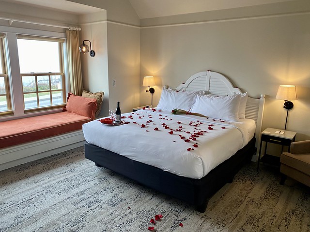 Saratoga Inn king hotel room with white bedding and red rose petals scattered on the king bed. A bottle of red Washington State wine, 2 wine glasses and heart-shaped box of chocolates are on the corner of the bed. A window seat provides a view of 2 sailboats docked in the small harbor across the street from the Washington State hotel. 
