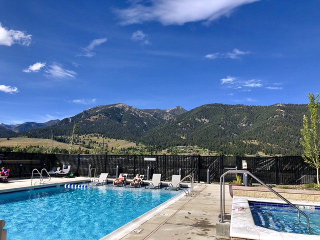 Two hotel guests lounge in the outdoor, heated pool at The Wilson Hotel Big Sky with Wilson Peak behind the pool. 